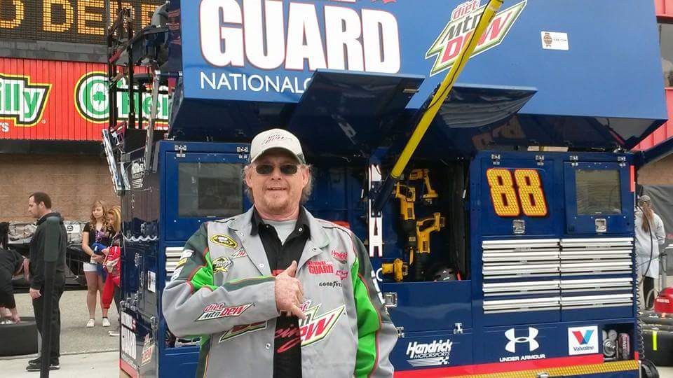 Bryan Carnett in a cap, sunglasses, and #88 crew gear giving a thumbs-up by the team hauler at a Cup race